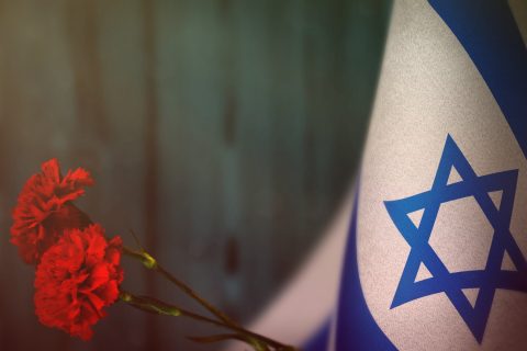 Israel Flag For Honour Of Veterans Day Or Memorial Day With Two Red Carnation Flowers Mockup. Glory To The Israel Heroes Of War Concept On Light Blue Blurred Natural Wood Wall. Background.